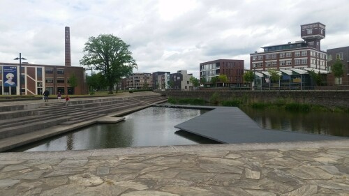 Der 13. Mai 2000 war ein schwarzer Tag in der niederländischen Stadt Enschede. Mitten im Stadtteil Roombeek explodierte die Feuerwerkfabrik S. E. Fireworks. 177 Tonnen Feuerwerkskörper legten das ganze Stadtquartier in Trümmer.

23 Menschen – darunter vier Feuerwehrmänner – starben, 947 Menschen wurden verletzt. 1.500 Wohngebäude wurden durch die Explosion beschädigt oder gerieten durch umherfliegende Feuerwerkskörper in Brand. 200 Wohnhäuser wurden vollständig zerstört und rund 300 als unbewohnbar erklärt. Darüber hinaus wurden über 100 Betriebe beschädigt. In nur einer Stunde verlieren mehr als 1.250 Einwohner ihr Zuhause.

Etwa zwei Drittel des 65 Hektar großen Stadtviertels, welches nördlich an den Hauptbahnhof und die Innenstadt anschließt, waren durch die Explosion betroffen. Mit der Katastrophe waren jedoch auch Chancen und Entwicklungspotenziale verbunden, welche einen Neuanfang für das durch die sterbende Textilindustrie geprägte Quartier bedeuten.

Der für den Wiederaufbau entwickelte Masterplan von Pi de Bruijn (de Architecten Cie. aus Amsterdam - www.cie.nl/projects/53 | Video: https://vimeo.com/51809482) verkörpert eine State of the Art-Stadtplanung. Die 1350 neu errichteten Wohnungen befinden sich in einem durchmischten Gebiet, welches durch kulturelle Angebote, Geschäfte und soziale Einrichtungen ergänzt wird. Freiräume, Wasser und Grün spielen ebenfalls eine große Rolle.

Dabei hat Pi de Bruijn den Einwohnern große Freiheit gelassen, selbst zu bestimmen, wie ihre Nachbarschaft aussehen soll. Üblicherweise macht die niederländische Stadtplanung vergleichsweise viele Vorgaben und lässt wenige Freiheiten. In Roombeek konnten rund 50 Prozent der Einwohner ihre neuen Wohngebäude vollkommen frei entwerfen, nur für die Bereiche an den Hauptverkehrsachsen wurden strengere Vorgaben gemacht. Das Straßennetz wurde exakt in der Form wieder aufgebaut, in der es vor der Explosion existierte. Dies sollte den ehemaligen Bewohnern ein rasches Ankommen und Einleben ermöglichen. Zudem wurde versucht, die vereinzelten noch stehenden Gebäude, soweit es geht, zu sichern und weiterzuentwickeln.