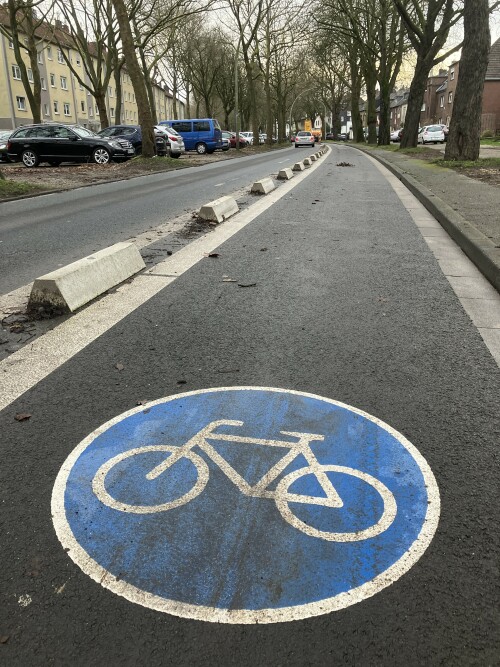 Geschützter Radfahrstreifen mit speziellen Betonelementen an der Hohenzollernstraße in Mönchengladbach. Am Beginn der PBL steht eine Warnbake, da die Trennelemente von Autofahrenden zu Beginn oft übersehen wurde. Am Knotenpunkt ist die PBL noch nicht durchgehend, der Radverkehr wird auf den Seitenbereich umgeleitet.